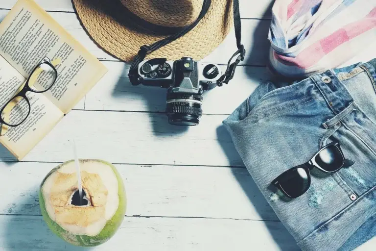 Flat lay of summer travel essentials including hat, camera, book, coconut drink, sunglasses, and clothes, representing a travel planning checklist for cheap flights and holidays.