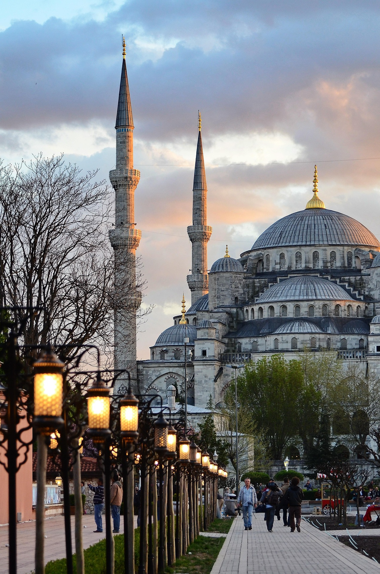 The historic Blue Mosque Sultan Ahmed Mosque in Istanbul at sunset with illuminated street lanterns and pedestrians on the walkway for tourists looking for the best hotels in Istanbul Sultanahmet