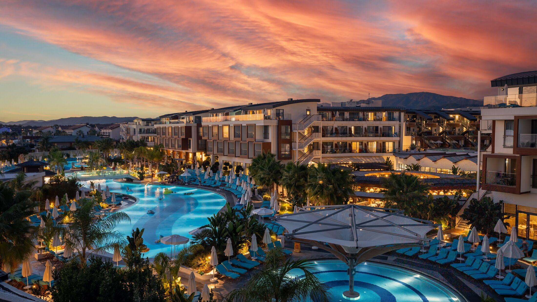 The luxury beachfront view of Liberty Fabay resort in Fethiye Turkey featuring large outdoor swimming pools with blue lounge chairs and palm trees along the Mediterranean coast at sunset