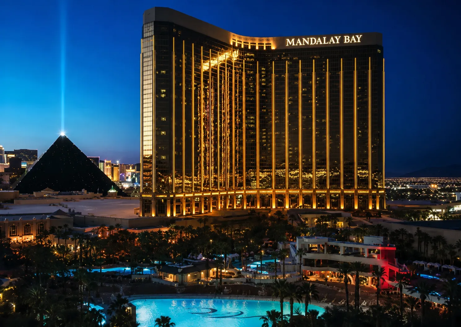 A high-angle night view of the Mandalay Bay Las Vegas resort, featuring its iconic golden, curved skyscraper illuminated with vertical lines of warm light. In the foreground, the extensive resort pool area and tropical landscaping are visible with glowing blue water. To the left, the dark silhouette of the Luxor pyramid stands against the twilight sky with its bright sky beam shooting straight up into the air.
